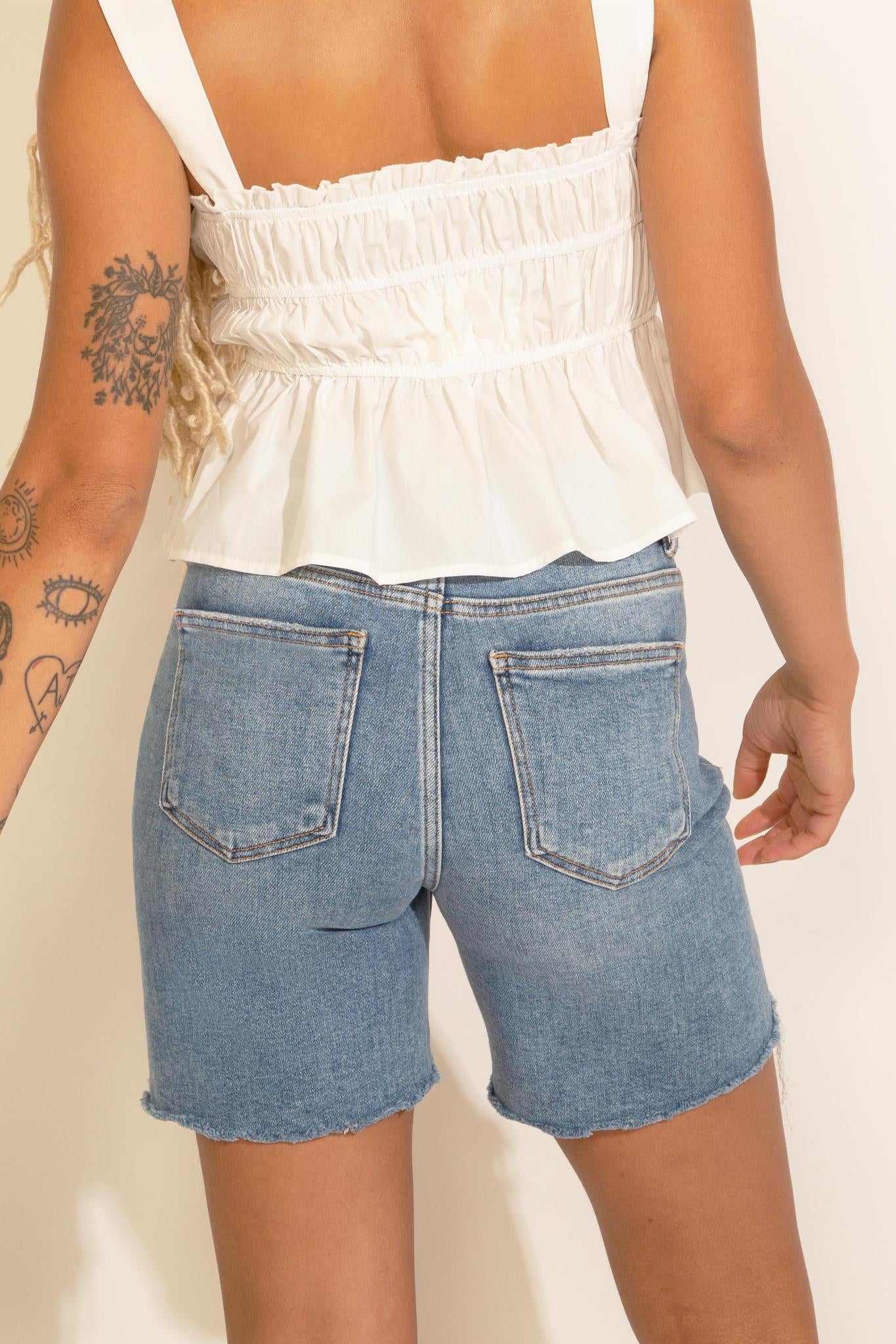 A woman wearing a sleeveless white peplum top with ruched detailing and an elastic banding, paired with blue denim shorts.