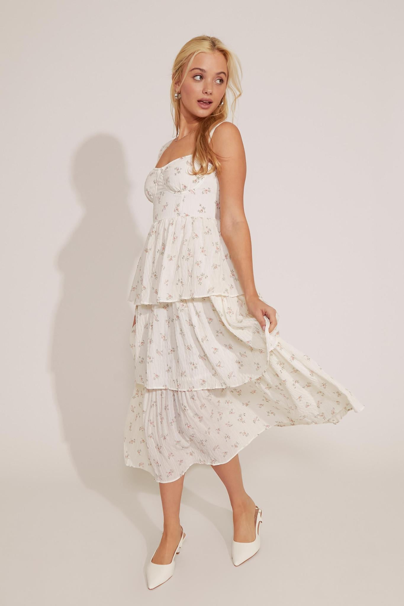A woman modeling a floral print midi dress with layered ruffles and bone wire structure, wearing white shoes.