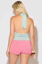 Model wearing a sage, flower detail, cowl halter neck, backless top with pink striped shorts on a white background.