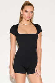 Model wearing a fitted black short-sleeve romper with a square neckline.