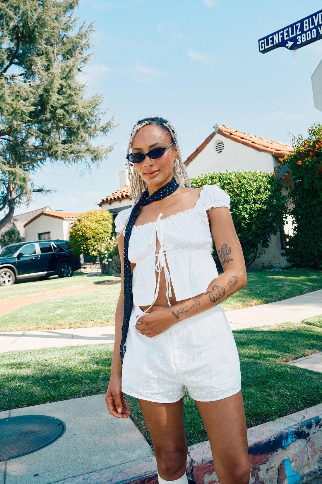 A woman wearing a white cap sleeve square neck open top with shorts, sunglasses, and a neck piece.