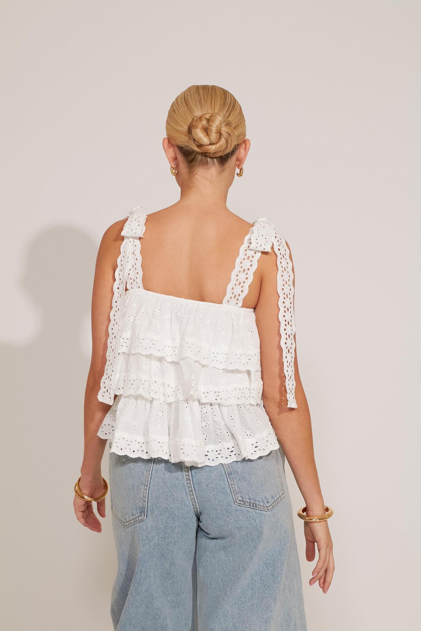 A woman sitting on the floor wearing a white tiered tank top with lace straps and floral embroidery, paired with blue denim shorts.