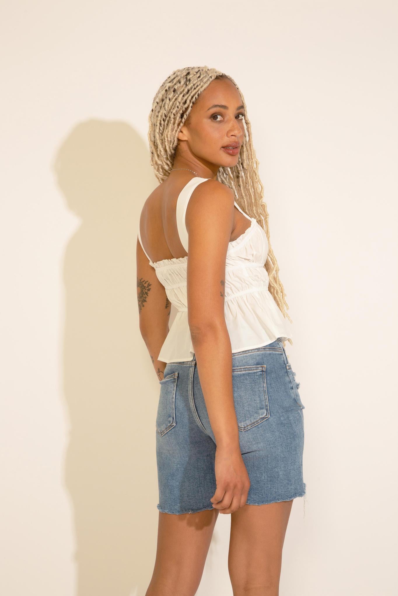 A woman wearing a sleeveless white peplum top with ruched detailing and an elastic banding, paired with blue denim shorts.