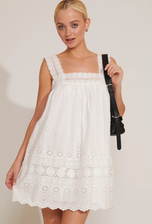 A woman wearing a white square-neck dress with eyelet detailing and shoulder straps. The dress features ruffle accents and a short length.
