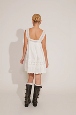 A woman wearing a white square-neck dress with eyelet detailing and shoulder straps. The dress features ruffle accents and a short length.