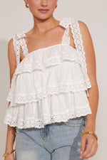 A woman sitting on the floor wearing a white tiered tank top with lace straps and floral embroidery, paired with blue denim shorts.