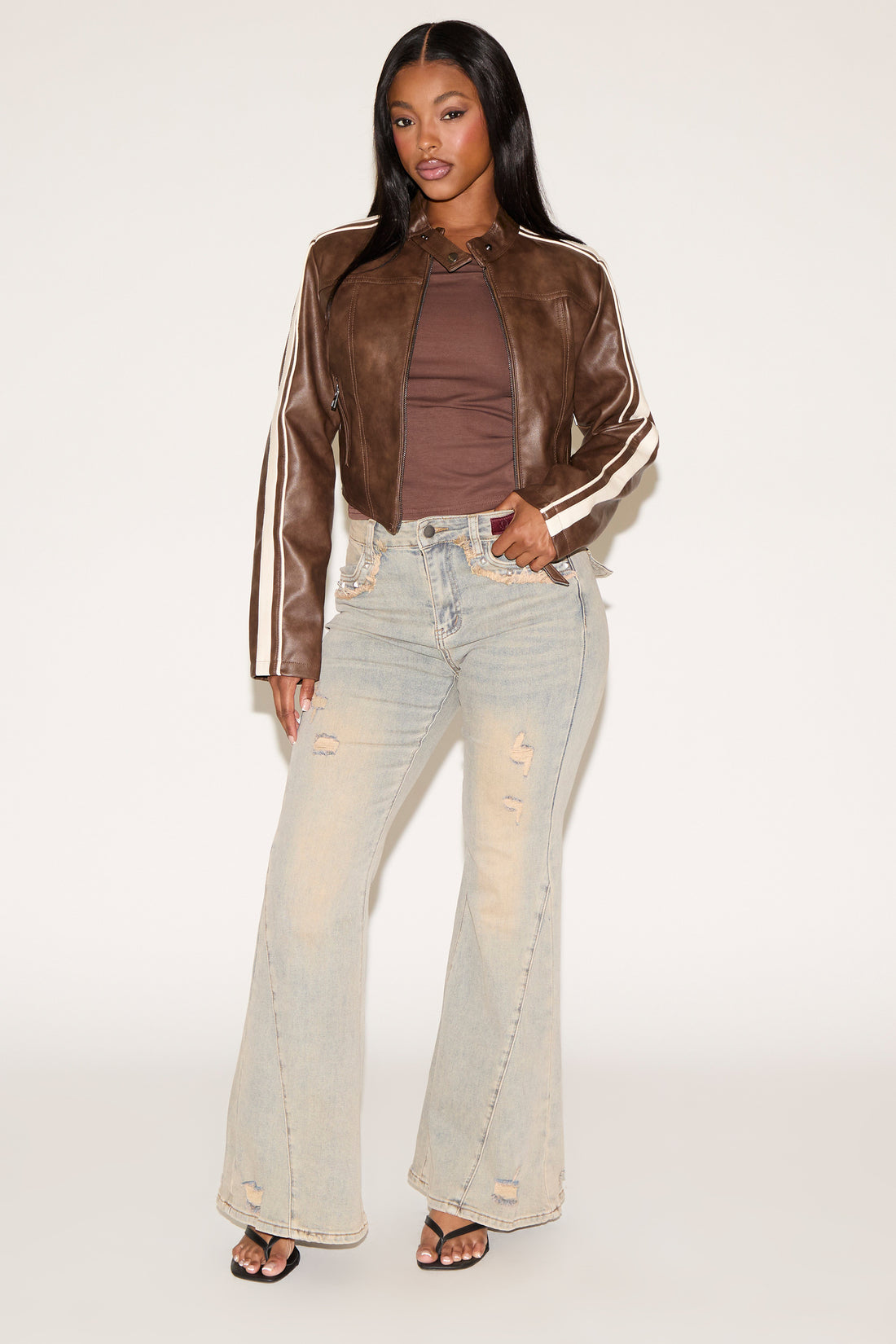 Woman wearing a brown leather jacket with white stripes, brown top, and light-colored jeans on a white background