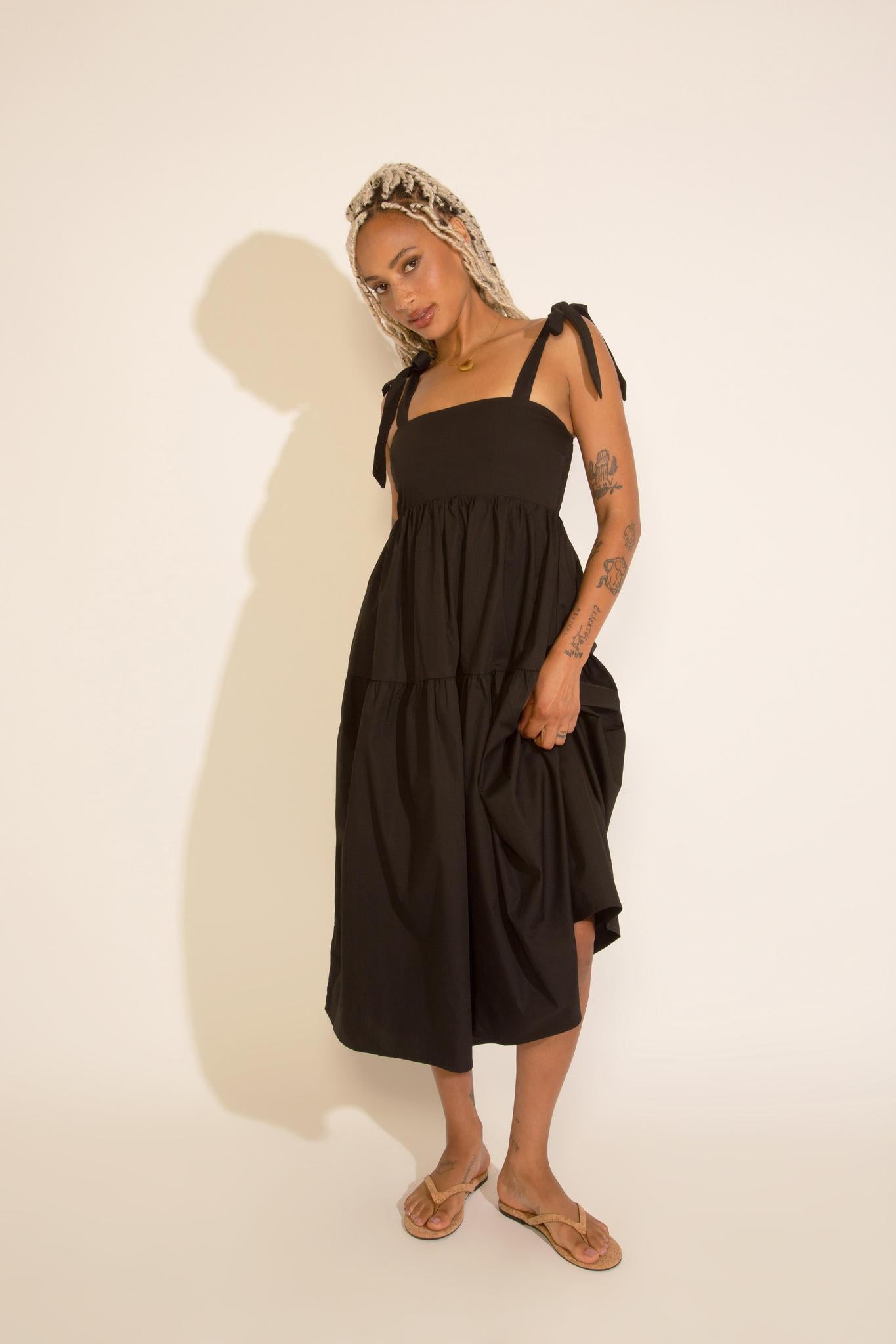 A woman standing wearing a black square neck midi dress with tied straps, a smocked bodice, and an A-line silhouette.