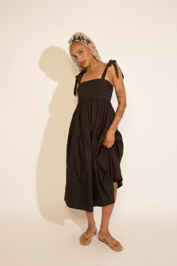 A woman standing wearing a black square neck midi dress with tied straps, a smocked bodice, and an A-line silhouette.