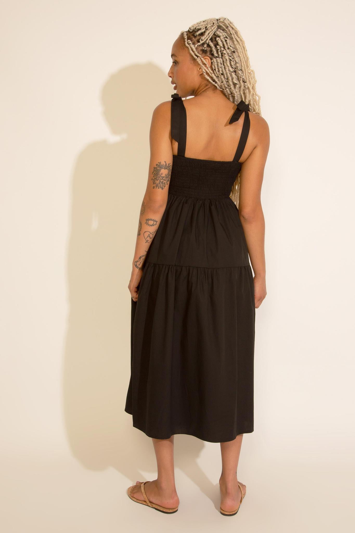 A woman standing wearing a black square neck midi dress with tied straps, a smocked bodice, and an A-line silhouette.