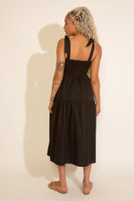 A woman standing wearing a black square neck midi dress with tied straps, a smocked bodice, and an A-line silhouette.