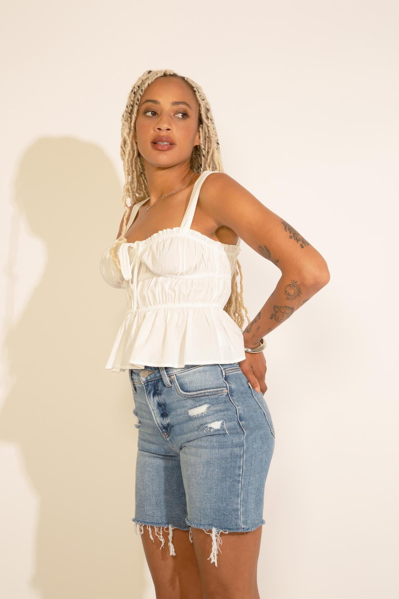 A woman wearing a sleeveless white peplum top with ruched detailing and an elastic banding, paired with blue denim shorts.