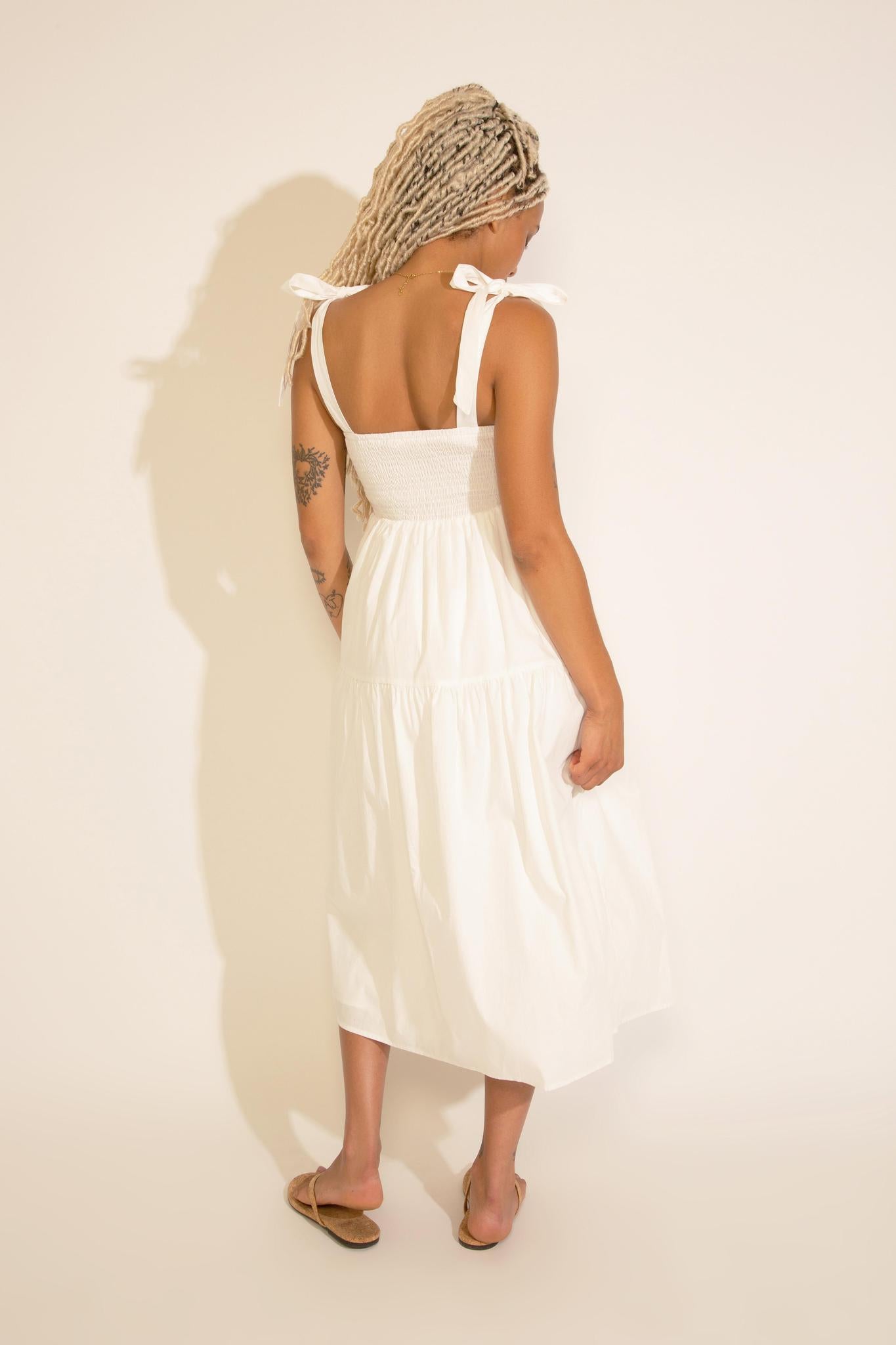 A woman standing wearing a white square neck midi dress with tied straps, a smocked bodice, and an A-line silhouette.