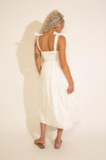 A woman standing wearing a white square neck midi dress with tied straps, a smocked bodice, and an A-line silhouette.
