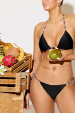 Woman in black bikini holding a green fruit with a wooden crate of fruits in the background