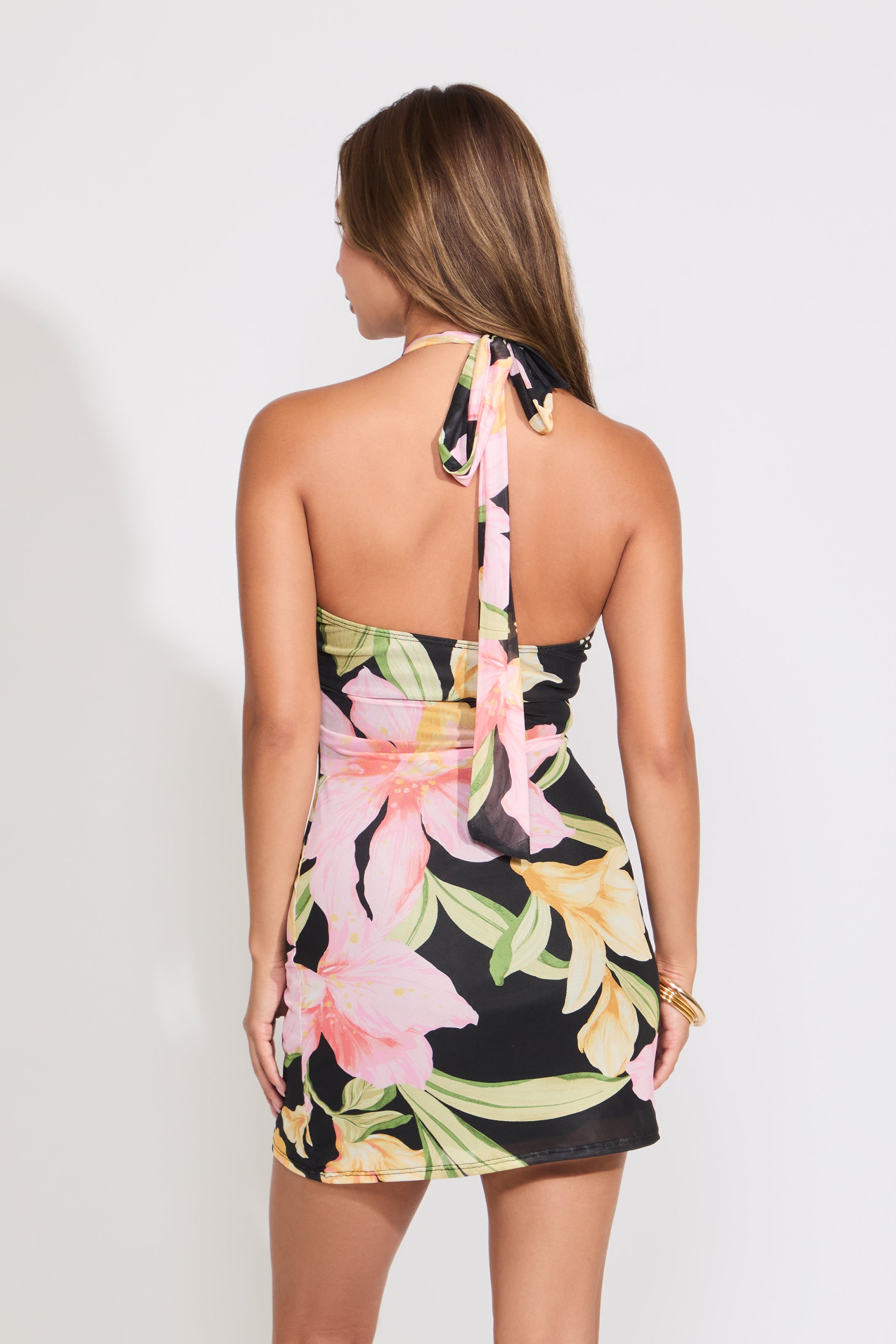 Model is wearing a floral tie neck mini dress with gold detail on  a white background. 
