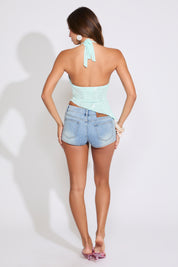 Model is wearing a mint SEQUINS EMBROIDERY ASYMMETRIC HEM TIE NECK TOP with light denim shorts on a white background.