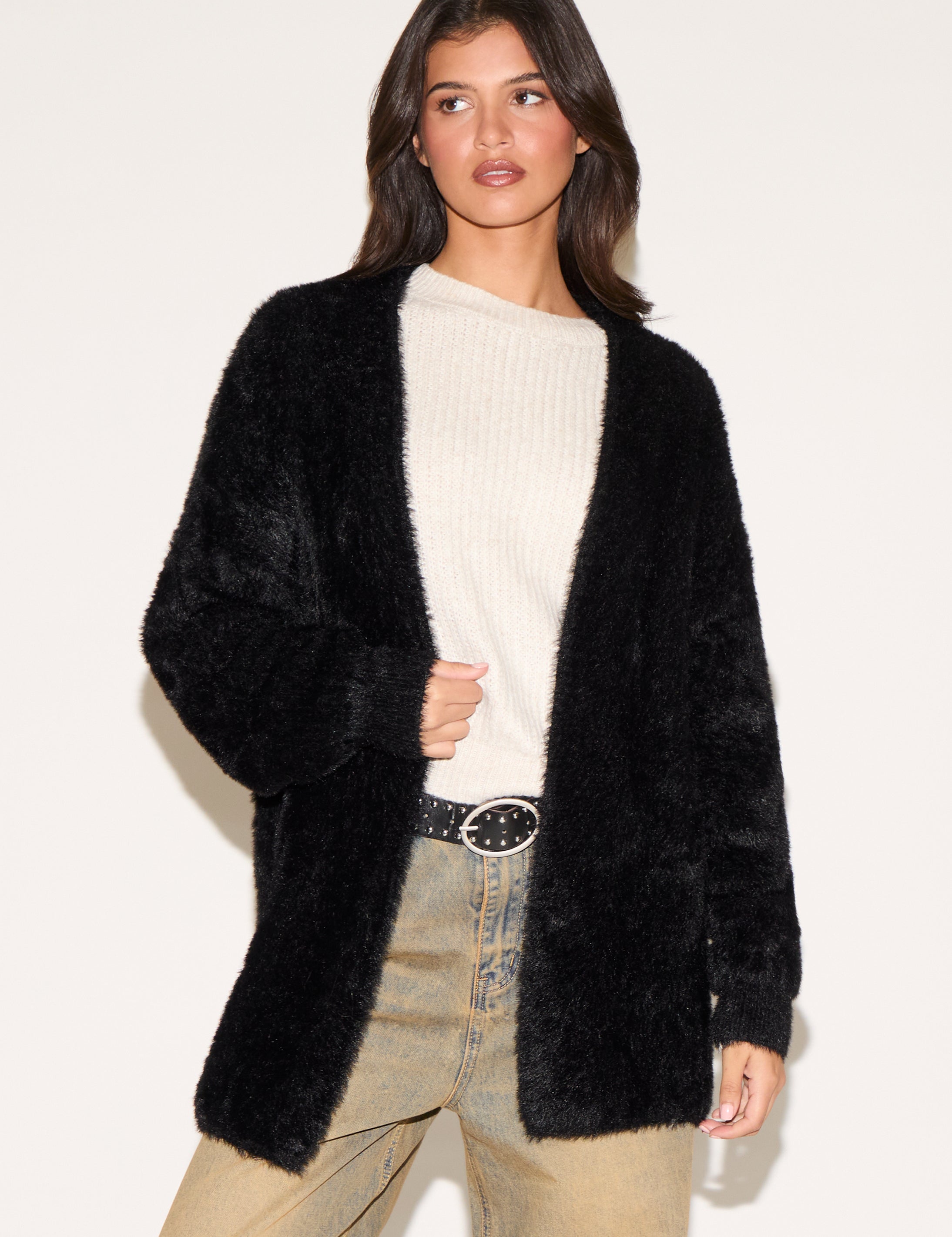 Woman wearing a black fluffy cardigan over a white sweater and beige pants on a light background