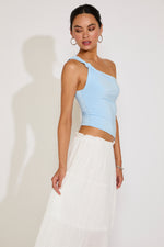 Woman wearing a light blue one-shoulder top and white pants against a plain background