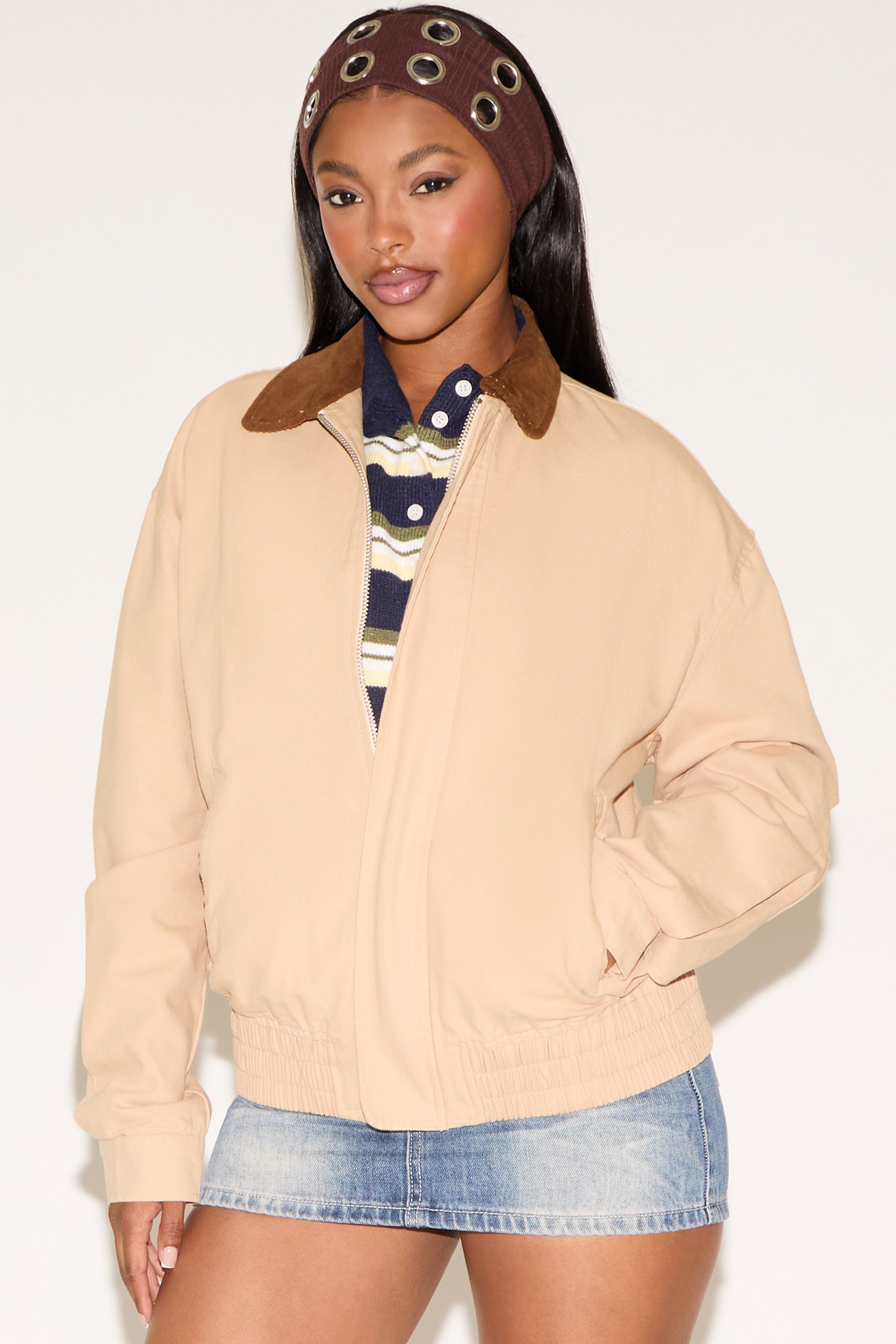 Woman wearing a beige jacket with a brown collar, striped shirt, and denim shorts on a white background
