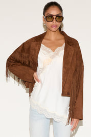 Woman wearing a brown suede jacket with fringe, white lace top, and light blue jeans against a white background.