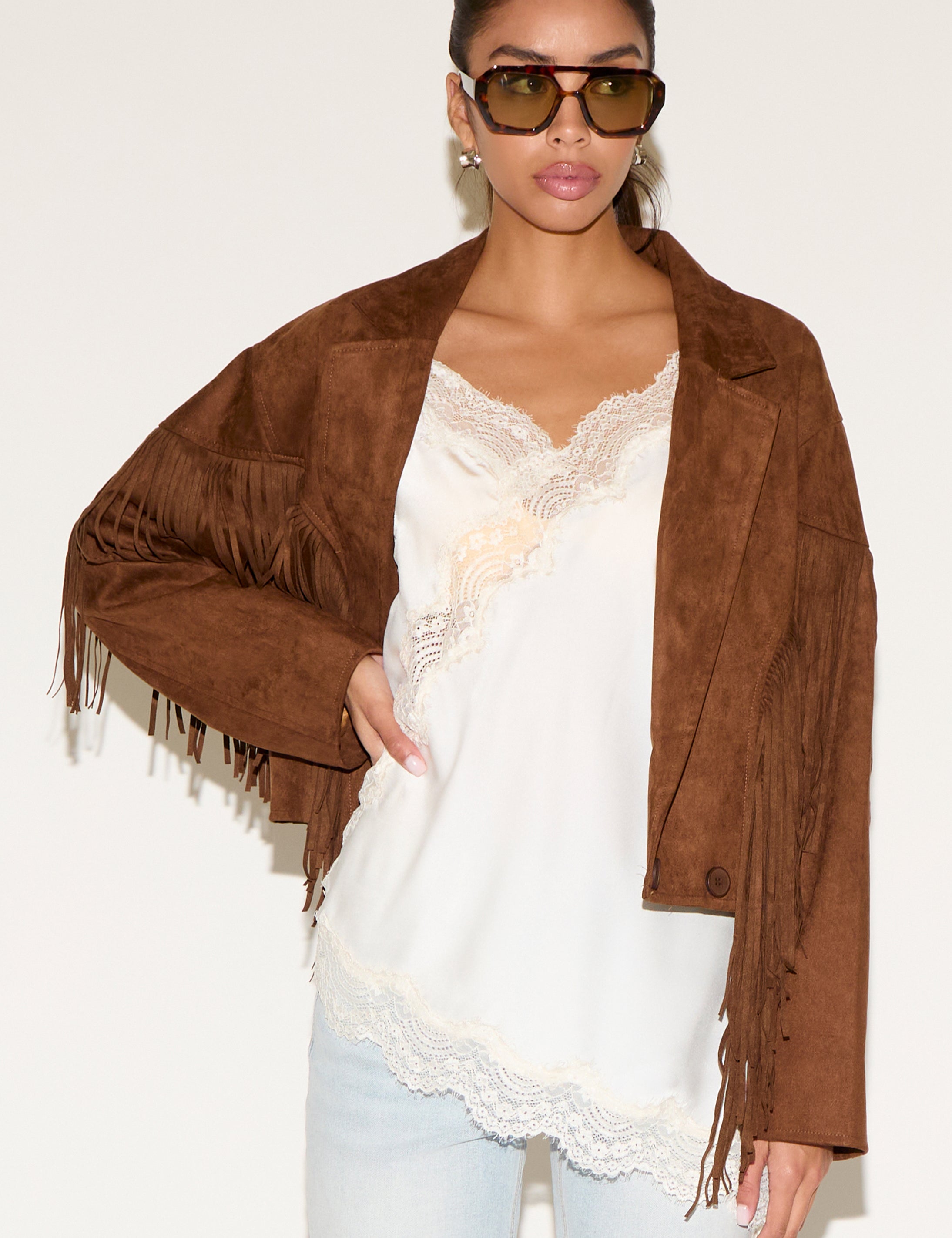 Woman wearing a brown suede jacket with fringe, white lace top, and light blue jeans against a white background.
