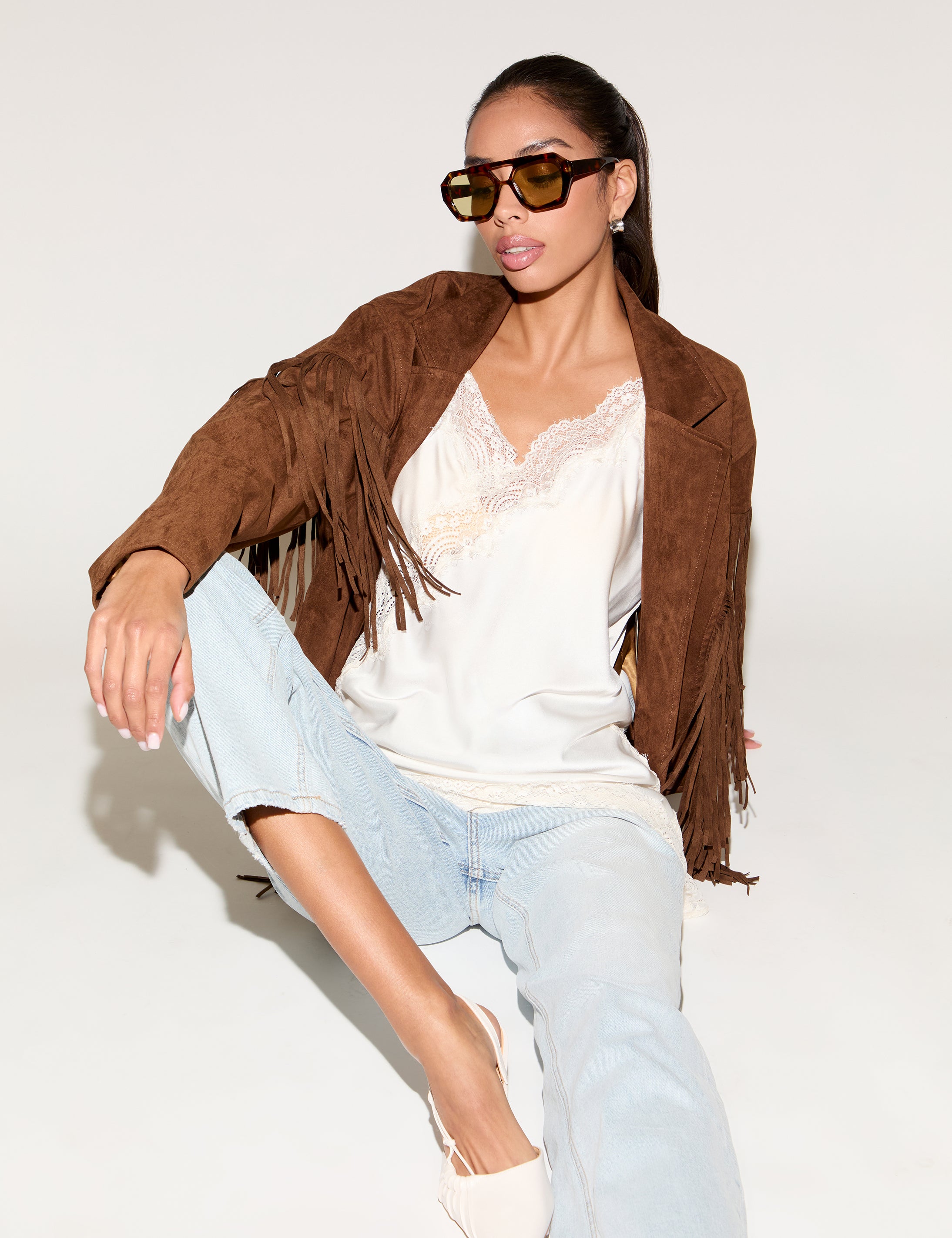 Woman wearing a brown suede jacket, white top, light blue jeans, and sunglasses on a white background