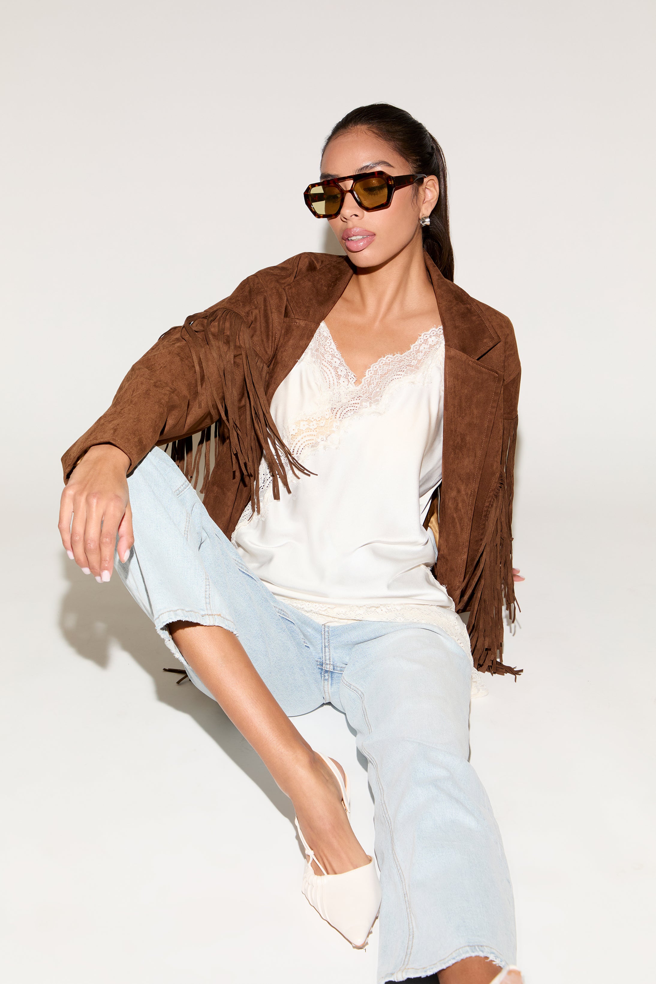 Woman wearing a brown suede jacket, white top, light blue jeans, and sunglasses on a white background