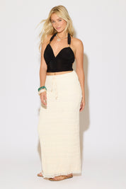 Model is wearing a black open back tie neck ruched top with metal detail with a cream maxi skirt on a white background.