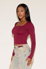 Woman wearing a burgundy long-sleeve top and light blue jeans against a white background