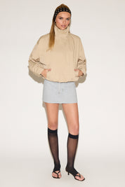 A woman models a beige zip-front
 jacket and a grey skirt against a white background.