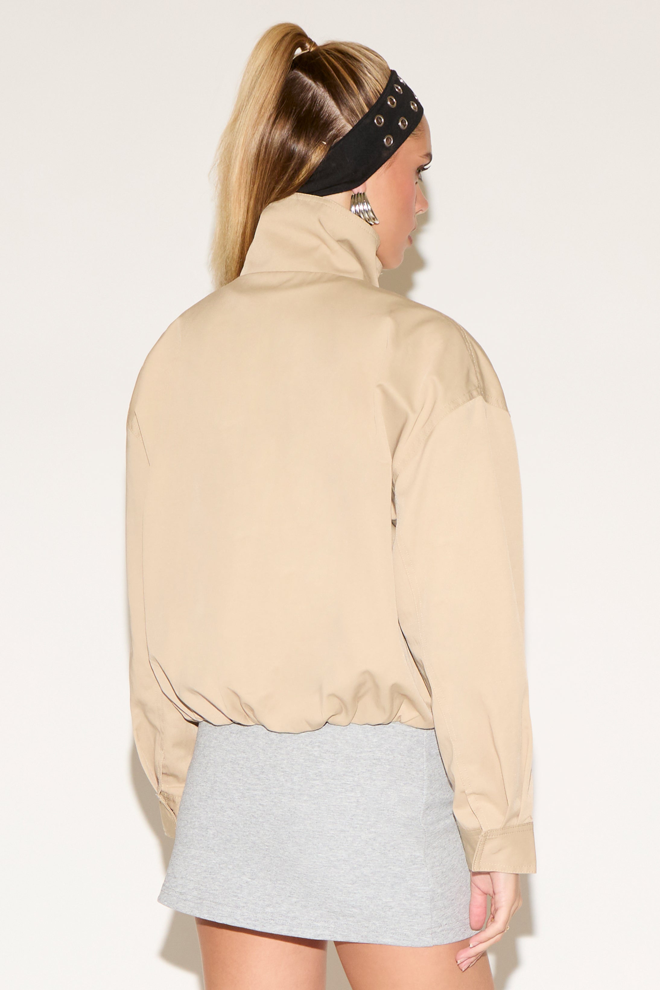 A woman models a beige zip-front jacke and a grey skirt against a white background.