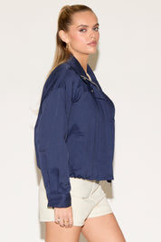 A woman models a navy blue zip-front jacket layered over a cream crop top and matching shorts against a white background