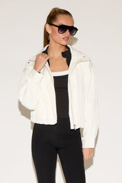 A woman models a navy blue zip-front jacket layered over a cream crop top and matching shorts against a white background.