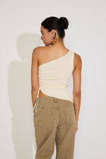 Woman wearing a beige one-shoulder top and beige pants against a white background