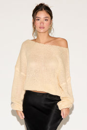 Woman wearing a beige off-shoulder sweater and black pants on a white background