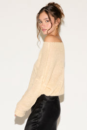 Woman wearing a cream off-shoulder sweater and black pants against a white background
