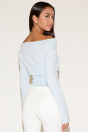 Woman wearing a light blue off-shoulder top and white pants on a plain background