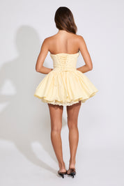 Model is wearing a yellow PLEATING DETAIL STRAPLESS RUFFLE HEM MINI DRESS with brown heels on a white background.