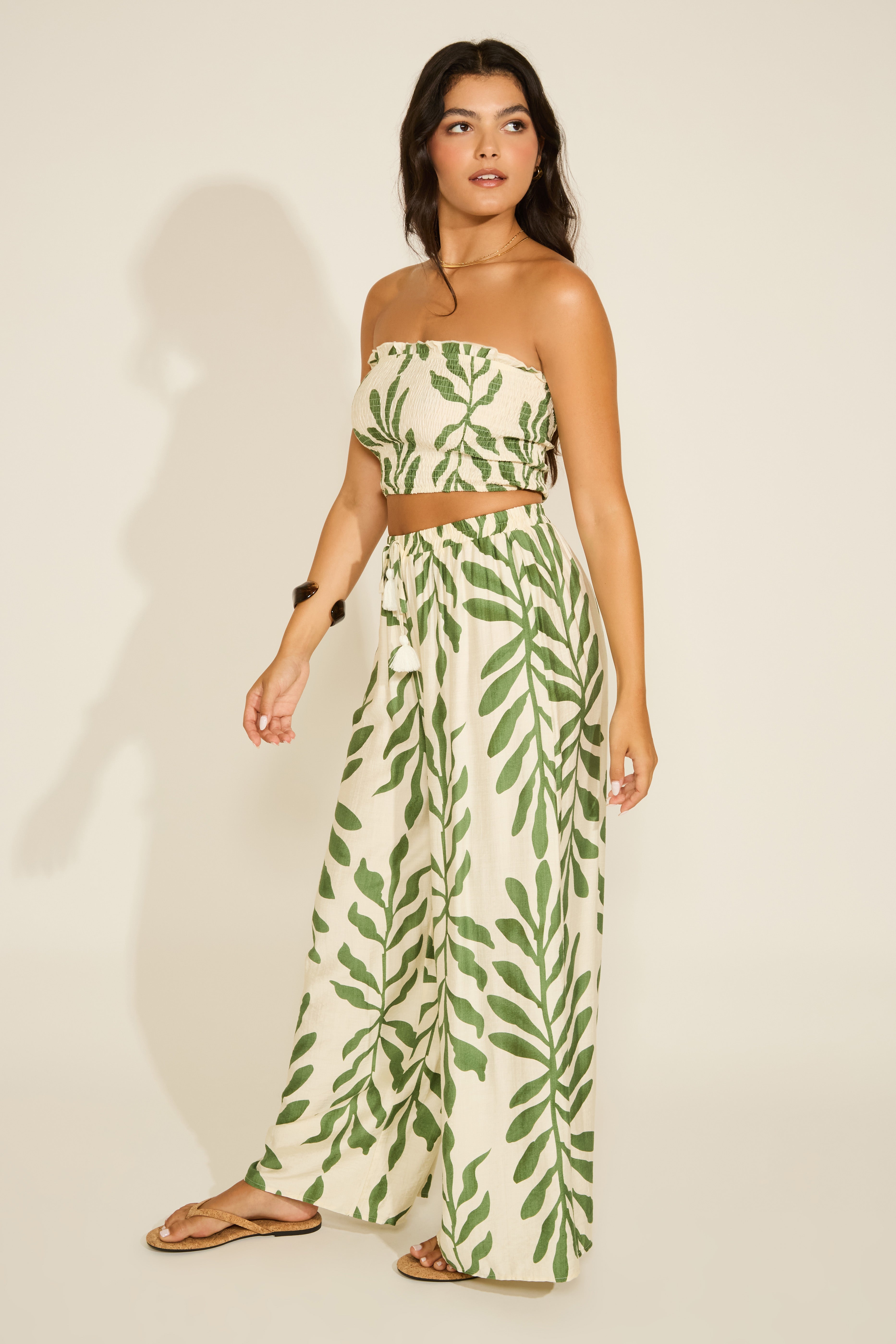 A model wearing a strapless leaf print smocked crop top and matching wide leg pants. The set is primarily in shades of green and white.