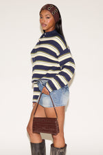 Woman wearing a blue, green and cream striped cardigan with a brown headband and denim blue skirt holding a brown purse on a plain white background