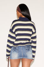Woman wearing a blue, green and cream striped cardigan with a brown headband and denim blue skirt holding a brown purse on a plain white background