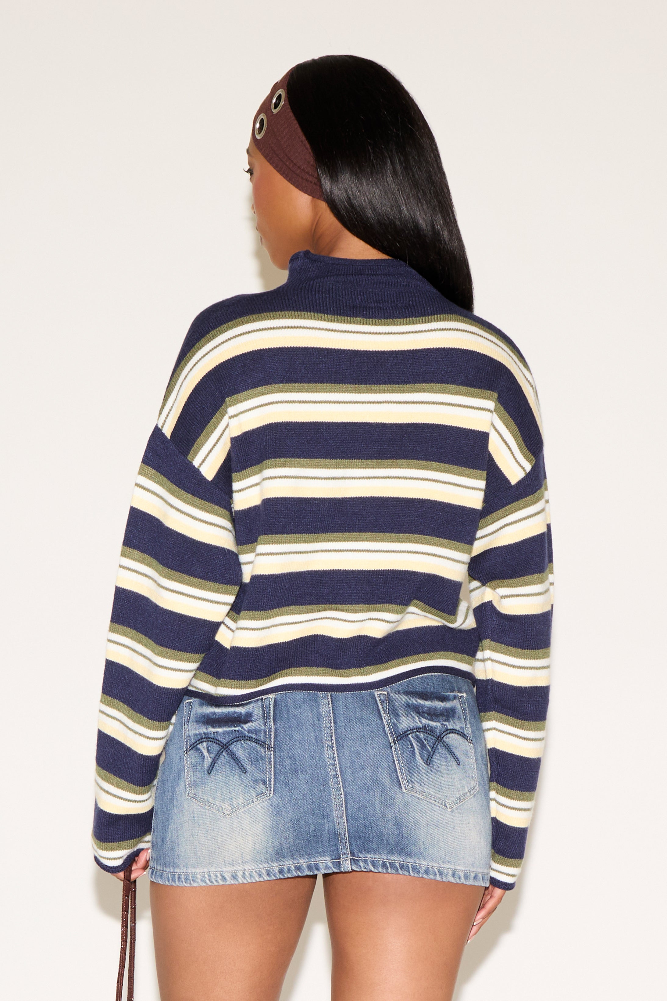 Woman wearing a blue, green and cream striped cardigan with a brown headband and denim blue skirt holding a brown purse on a plain white background