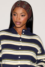 Woman wearing a blue, green and cream striped cardigan with a brown headband and denim blue skirt holding a brown purse on a plain white background