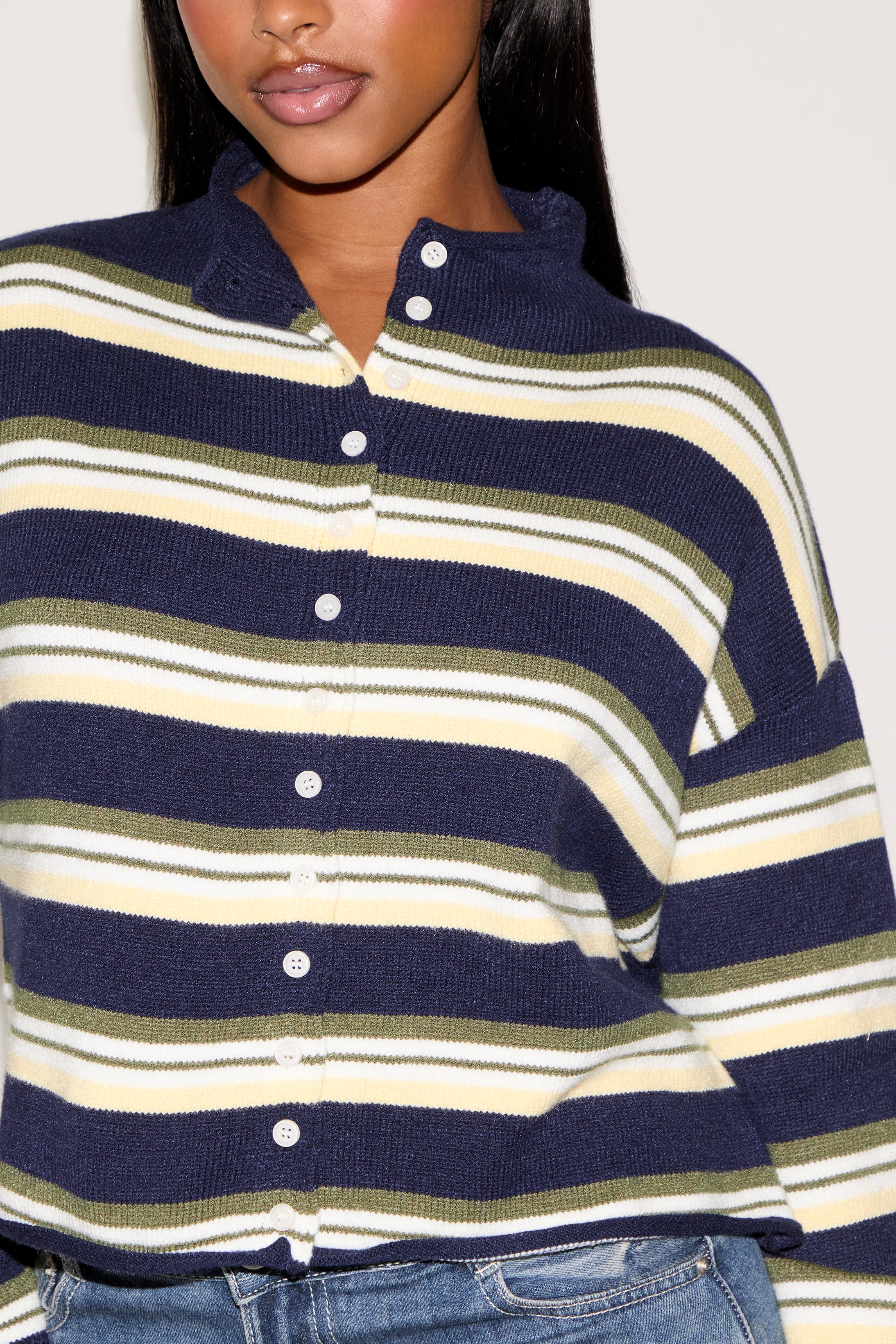Woman wearing a blue, green and cream striped cardigan with a brown headband and denim blue skirt holding a brown purse on a plain white background