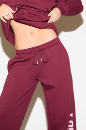 A woman poses against a white background wearing a burgundy cropped hoodie and matching wide-leg sweatpants with “Los Angeles” text down one leg.
