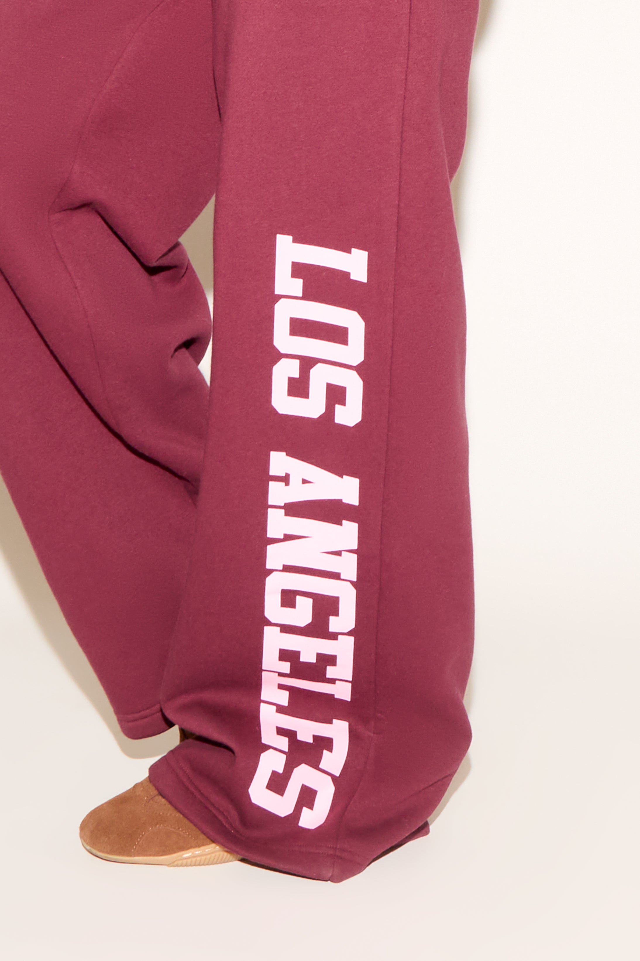 A woman poses against a white background wearing a burgundy cropped hoodie and matching wide-leg sweatpants with “Los Angeles” text down one leg.