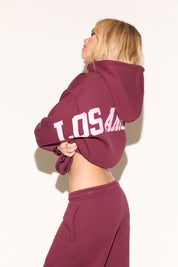 A woman poses against a white background wearing a burgundy cropped hoodie and matching wide-leg sweatpants. 