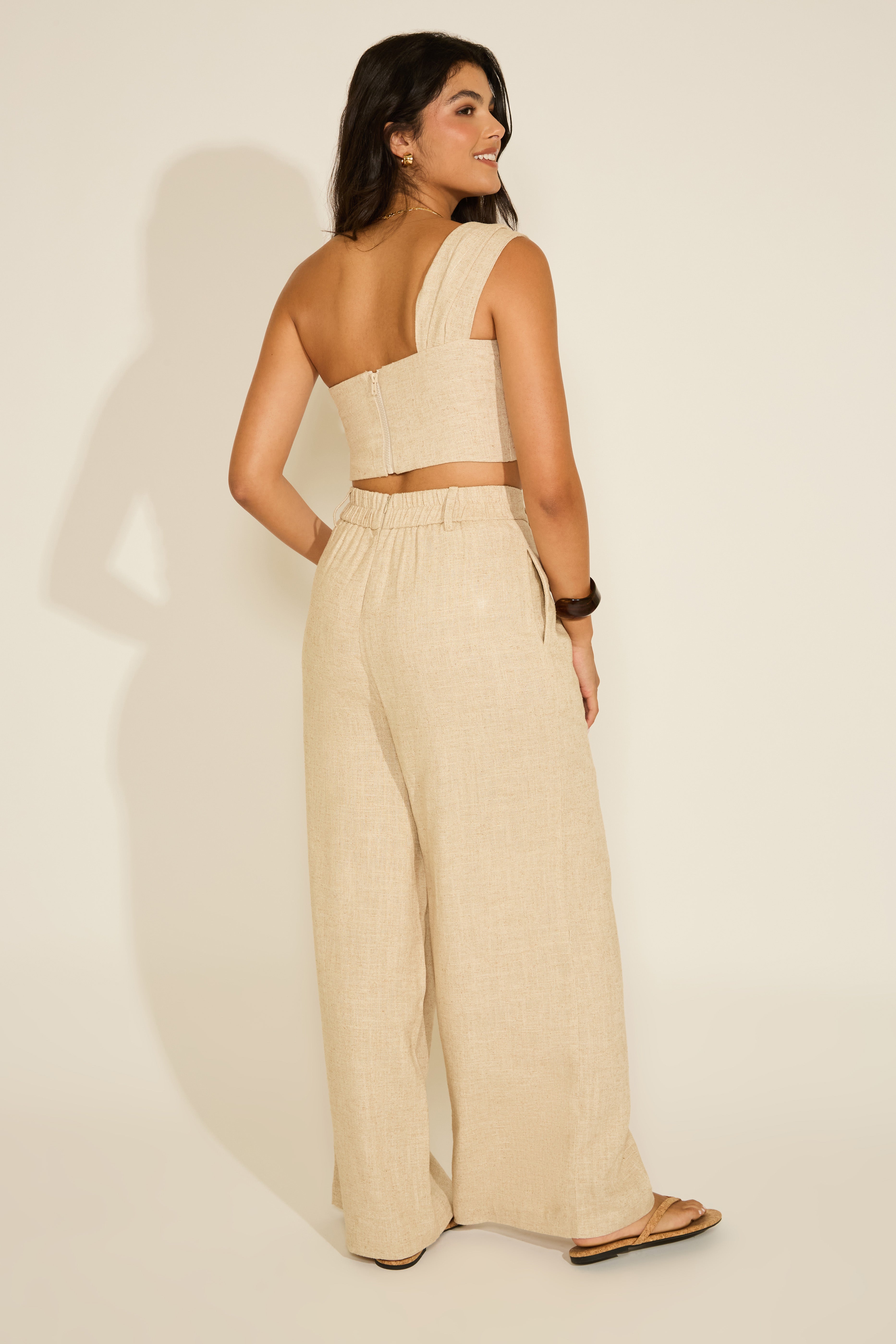 Woman wearing a beige one-shoulder top and wide-leg pants against a plain background.