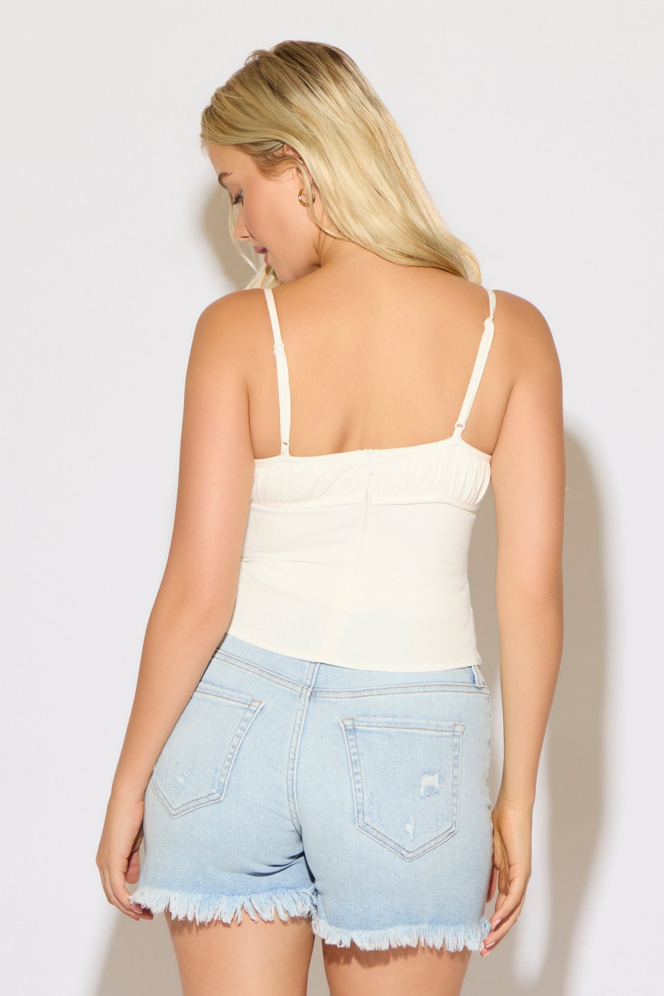Model is wearing a white ruched bust strappy cami top with denim shorts on a white background. 